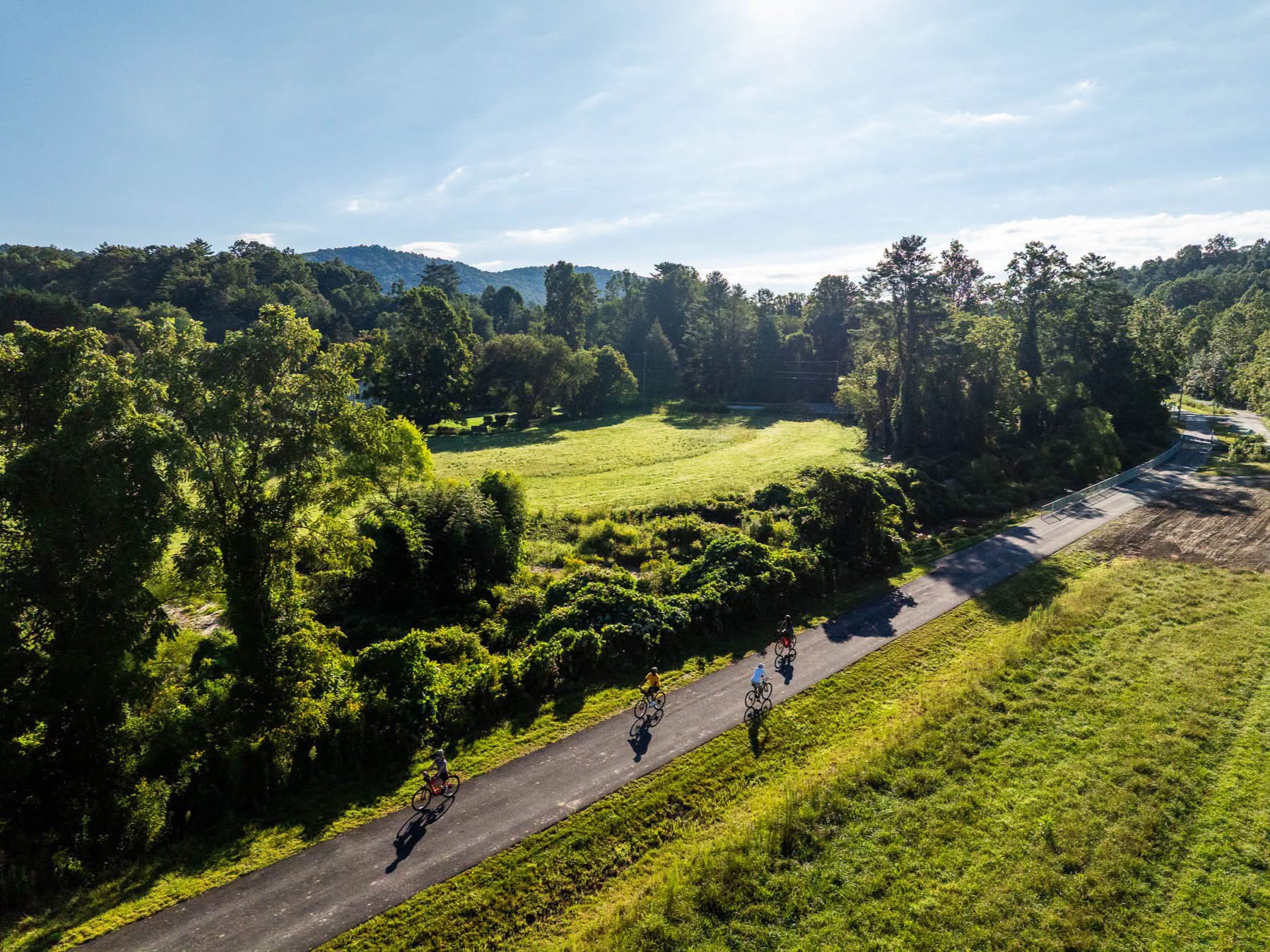 Cyclists riding the paved Ecusta Trail through green fields with mountains in the distance